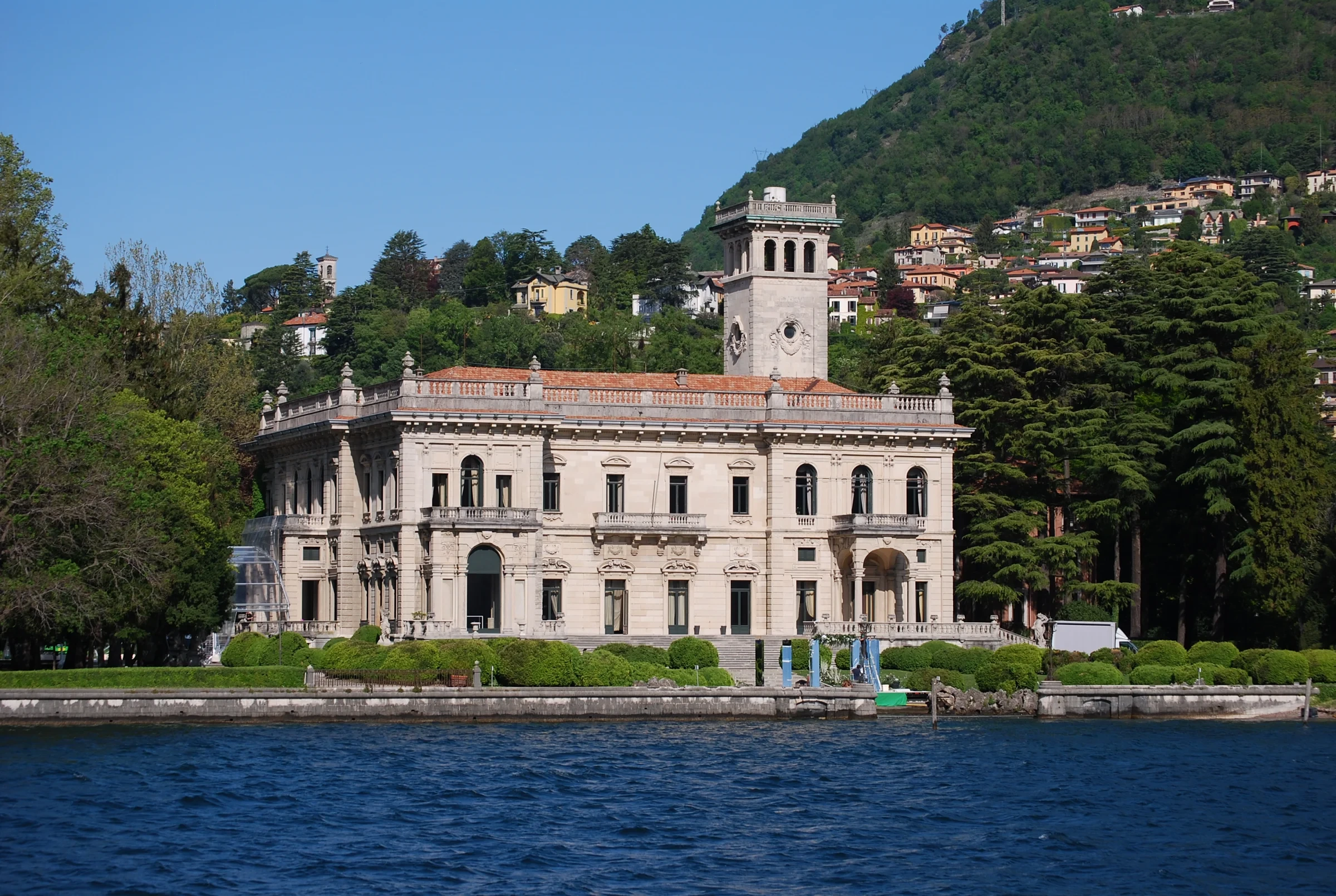 Villa Erba’s pale façade and tower beside Lake Como, with a wooded hillside behind.