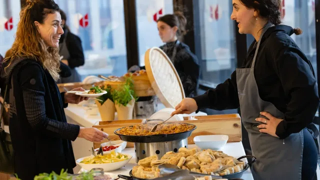 A staff member serves food from a pan while speaking with a guest holding a plate at a buffet.