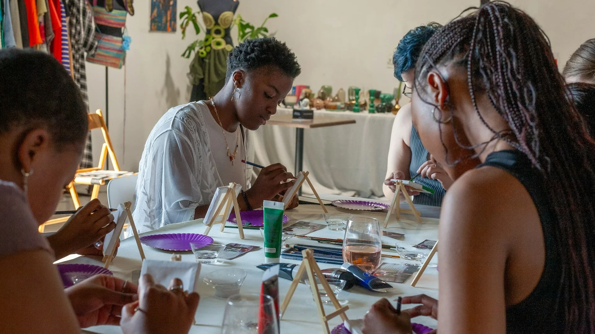 Deelnemers schilderen kleine doeken aan een gezamenlijke workshoptafel met penselen, paletten en ezels.