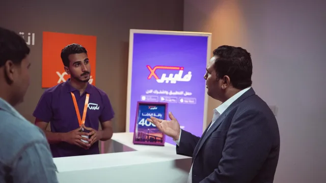 A visitor gestures across an exhibition counter while a staff member in a purple shirt listens.