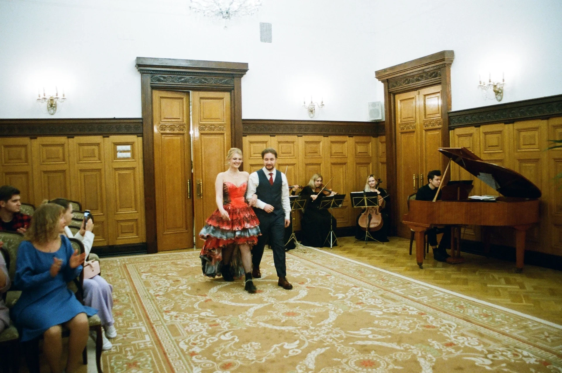 Violinist, cellist and pianist performing behind a formally dressed couple in a wood-panelled hall with seated guests