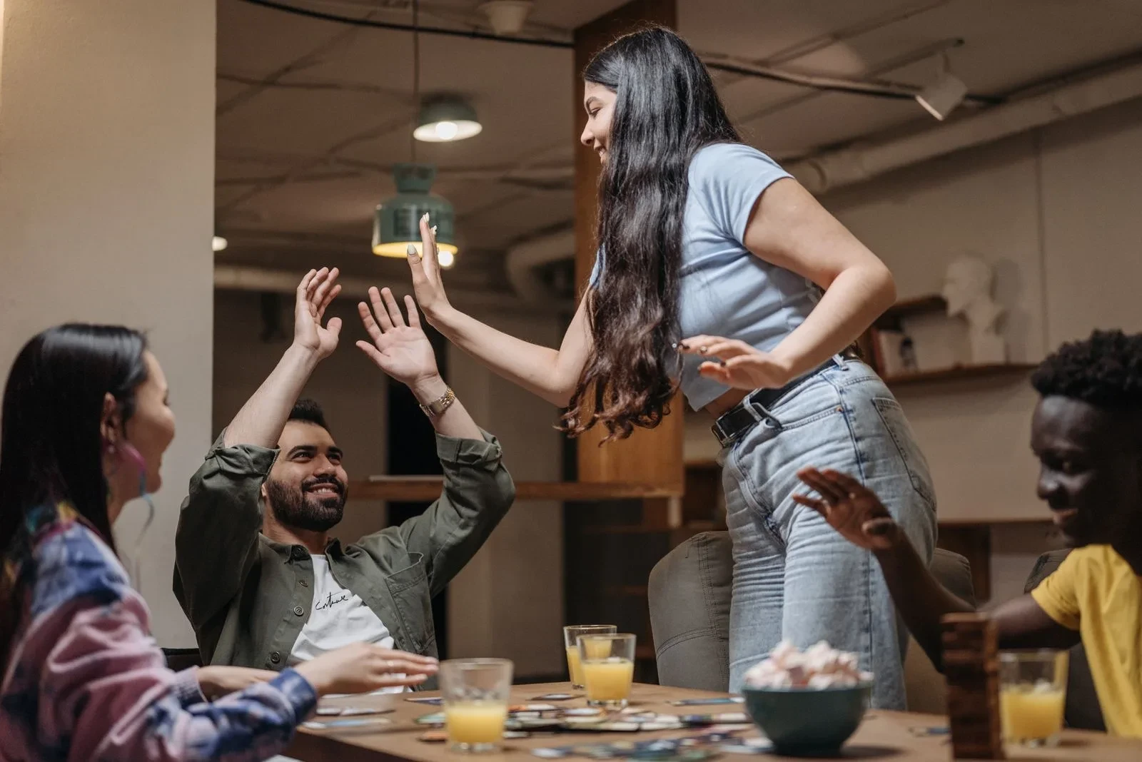 Des amis lèvent les mains autour d’une table avec des pièces de jeu et des boissons.