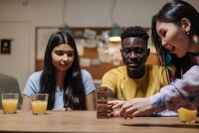 Drie mensen aan een tafel, van wie één voorzichtig een blok uit een houten stapeltoren haalt.