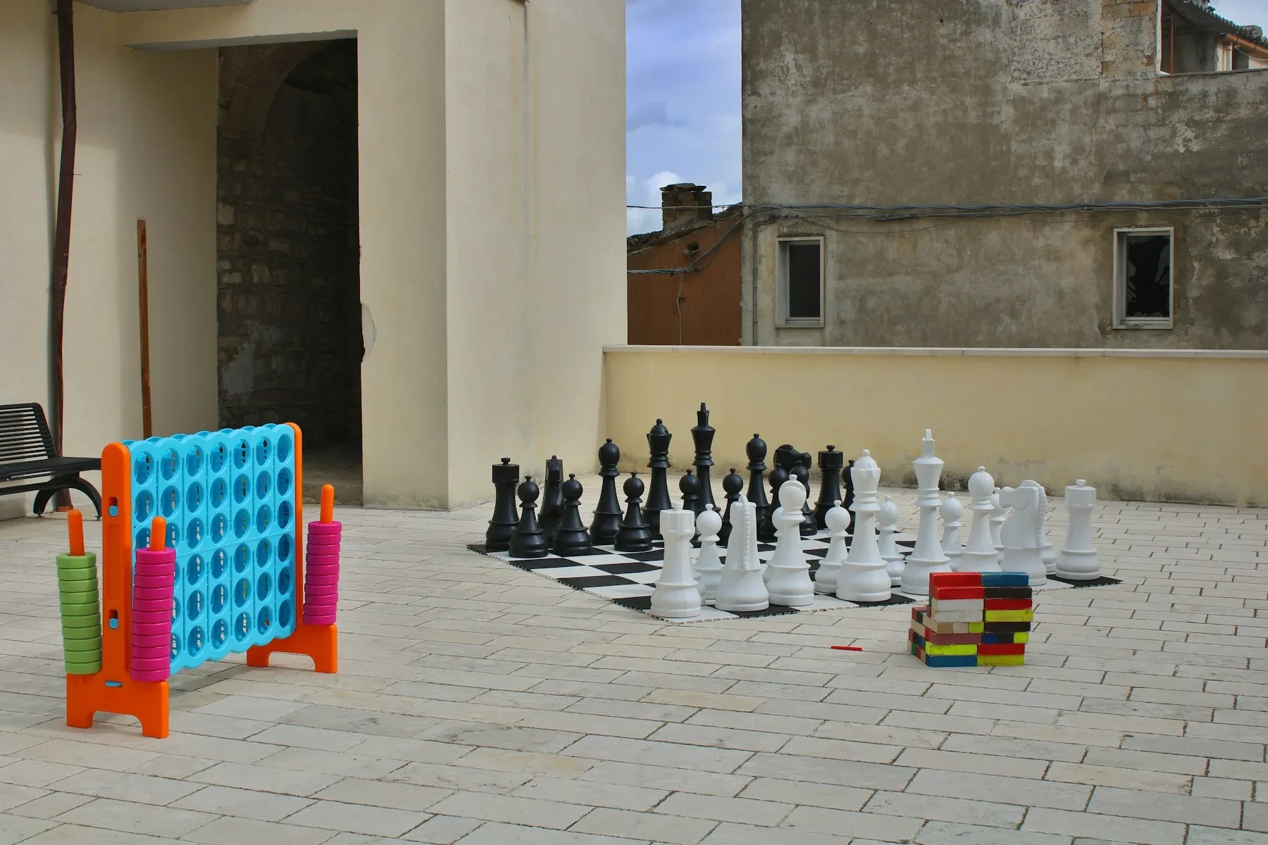 Un quatre-à-la-suite géant coloré, de grandes pièces d’échecs et des blocs en bois sur une terrasse pavée.