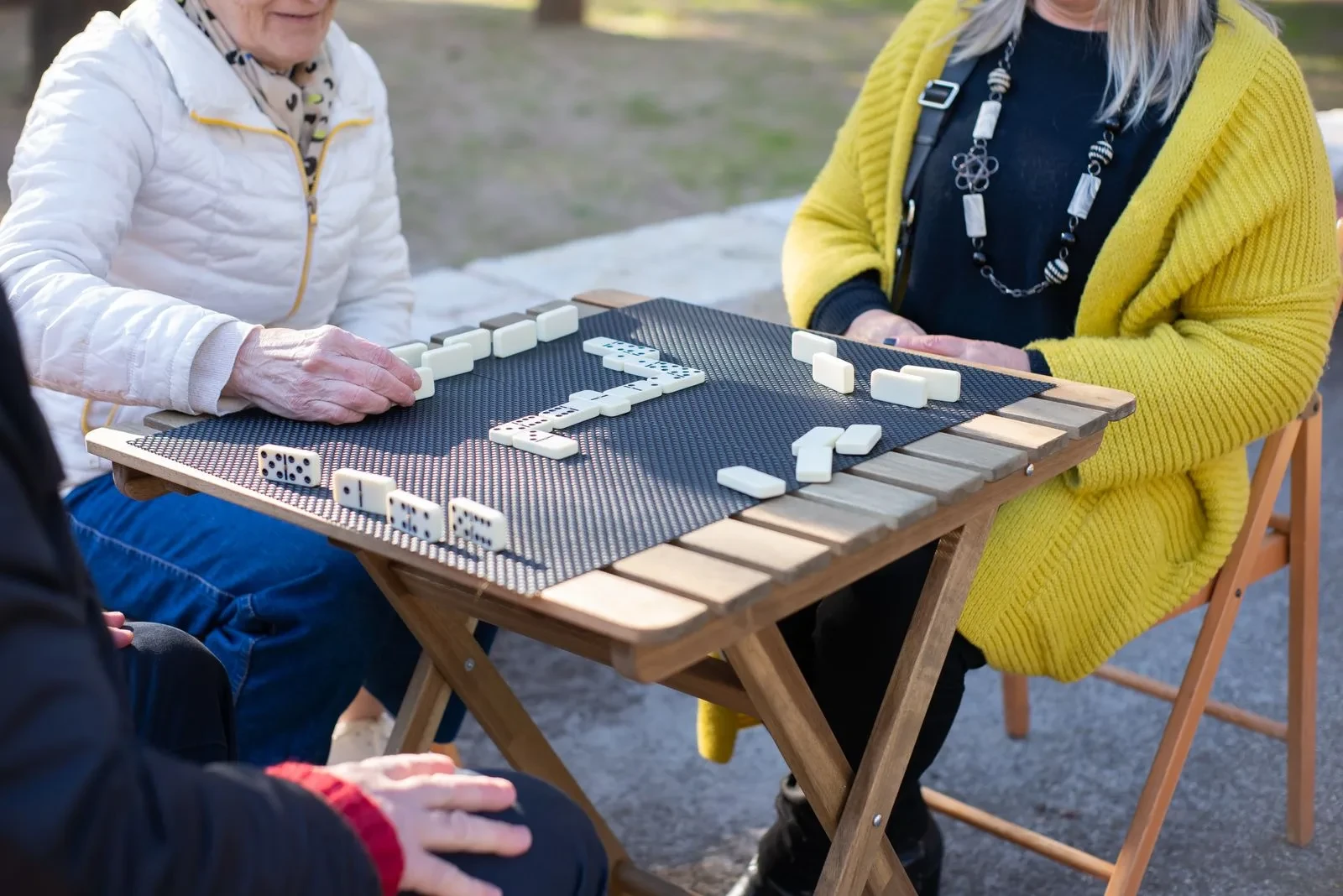 Des adultes âgés jouent aux dominos autour d’une table en bois à l’extérieur.
