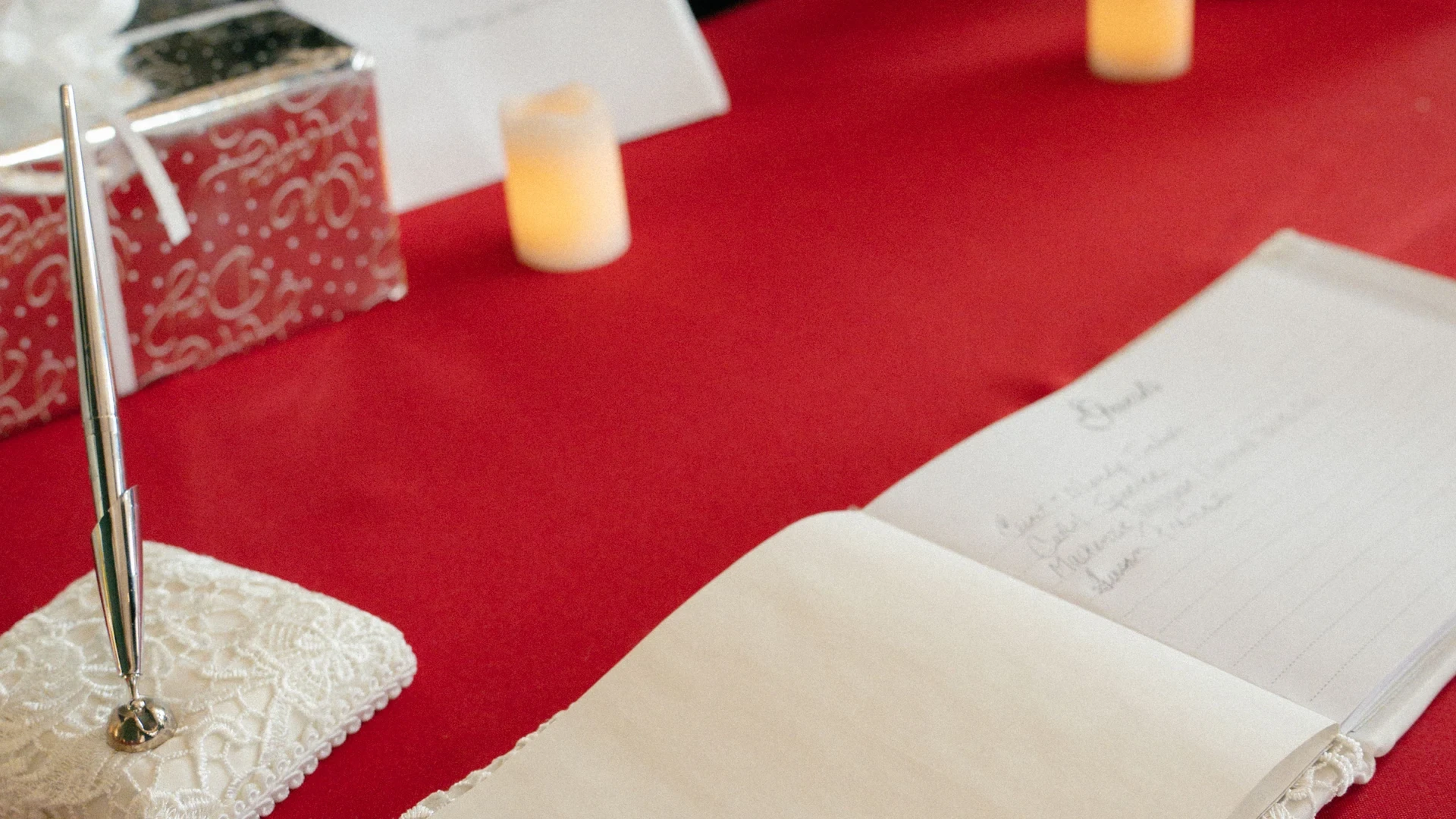 An open guest book with handwritten entries and a silver pen on a red reception tablecloth.