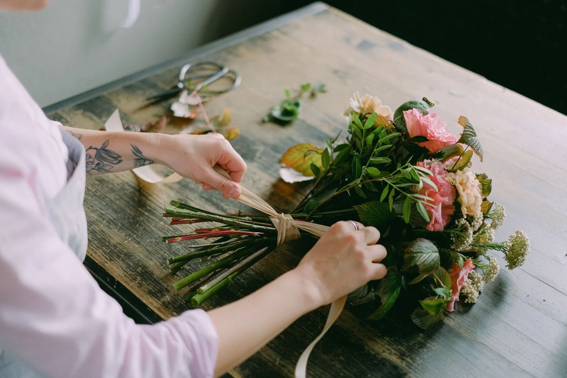 A person ties a pale ribbon around the stems of a pink and white bouquet on a wooden worktable.
