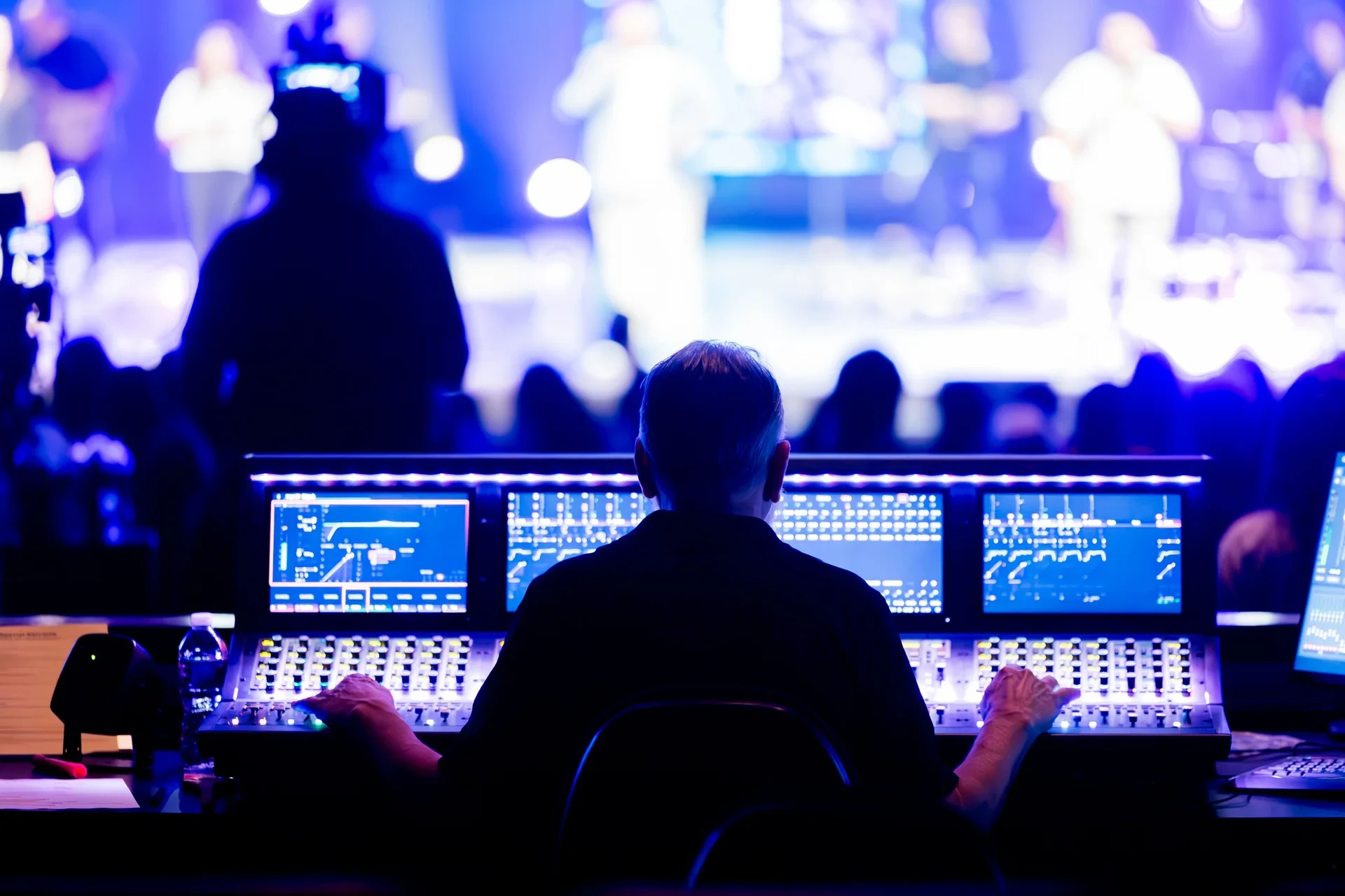A sound engineer operates a digital mixing console facing a live performance and its audience.