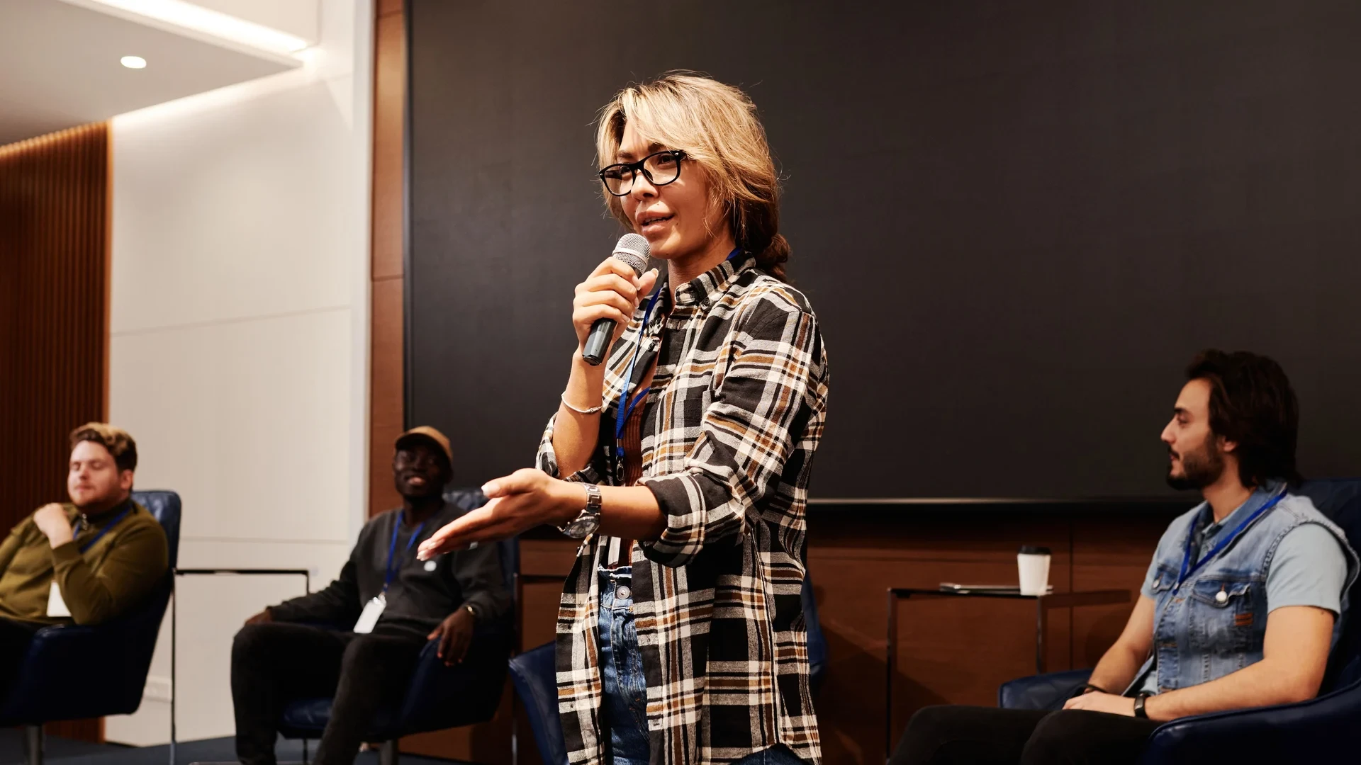A woman speaks into a handheld microphone in front of seated panel participants.