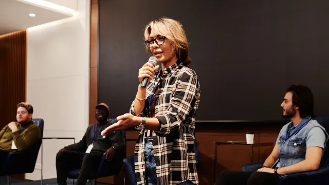 Een vrouw spreekt in een handmicrofoon voor zittende paneldeelnemers.