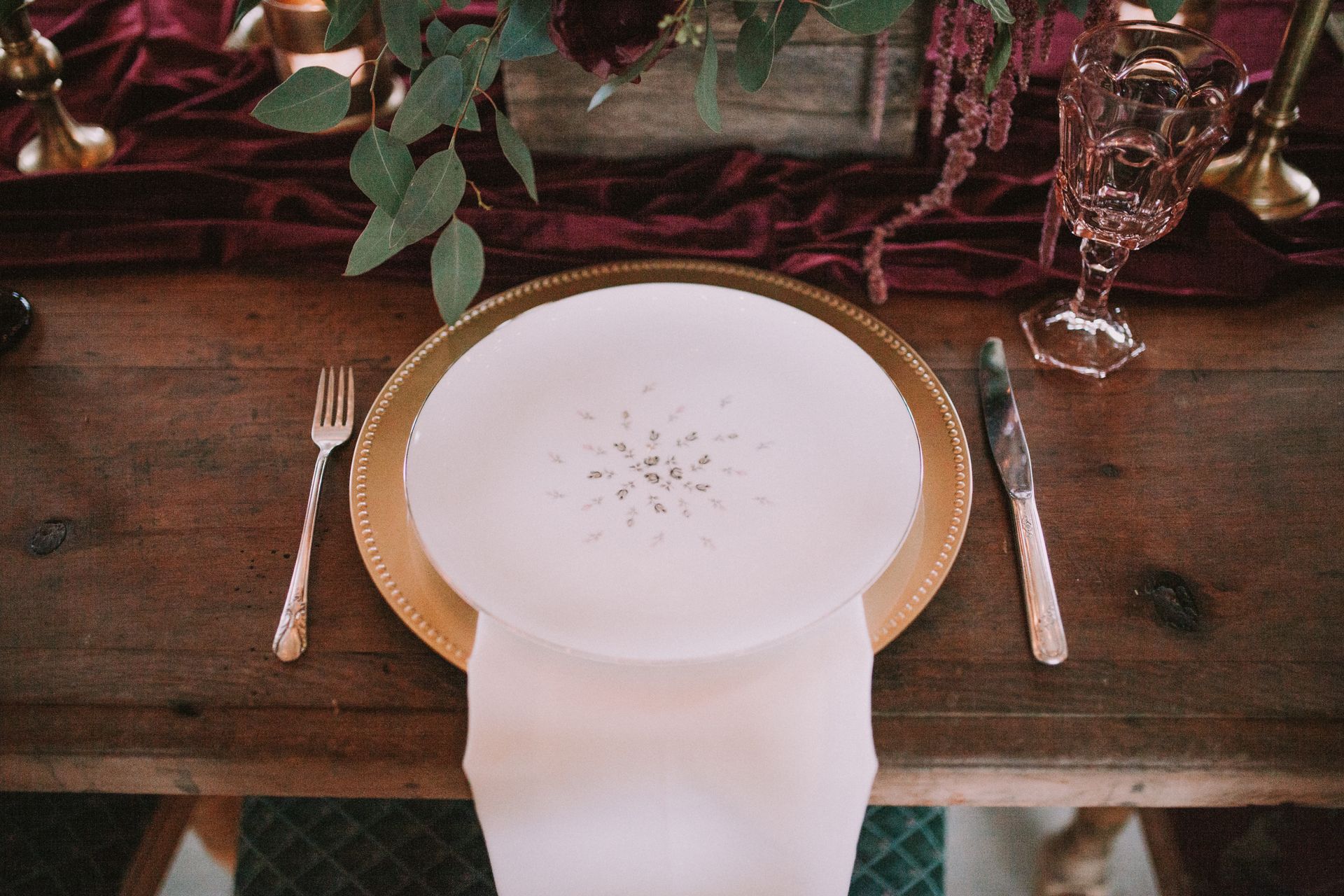 Pale plate and cream napkin on a gold-toned charger, with burgundy fabric, greenery and pink glassware