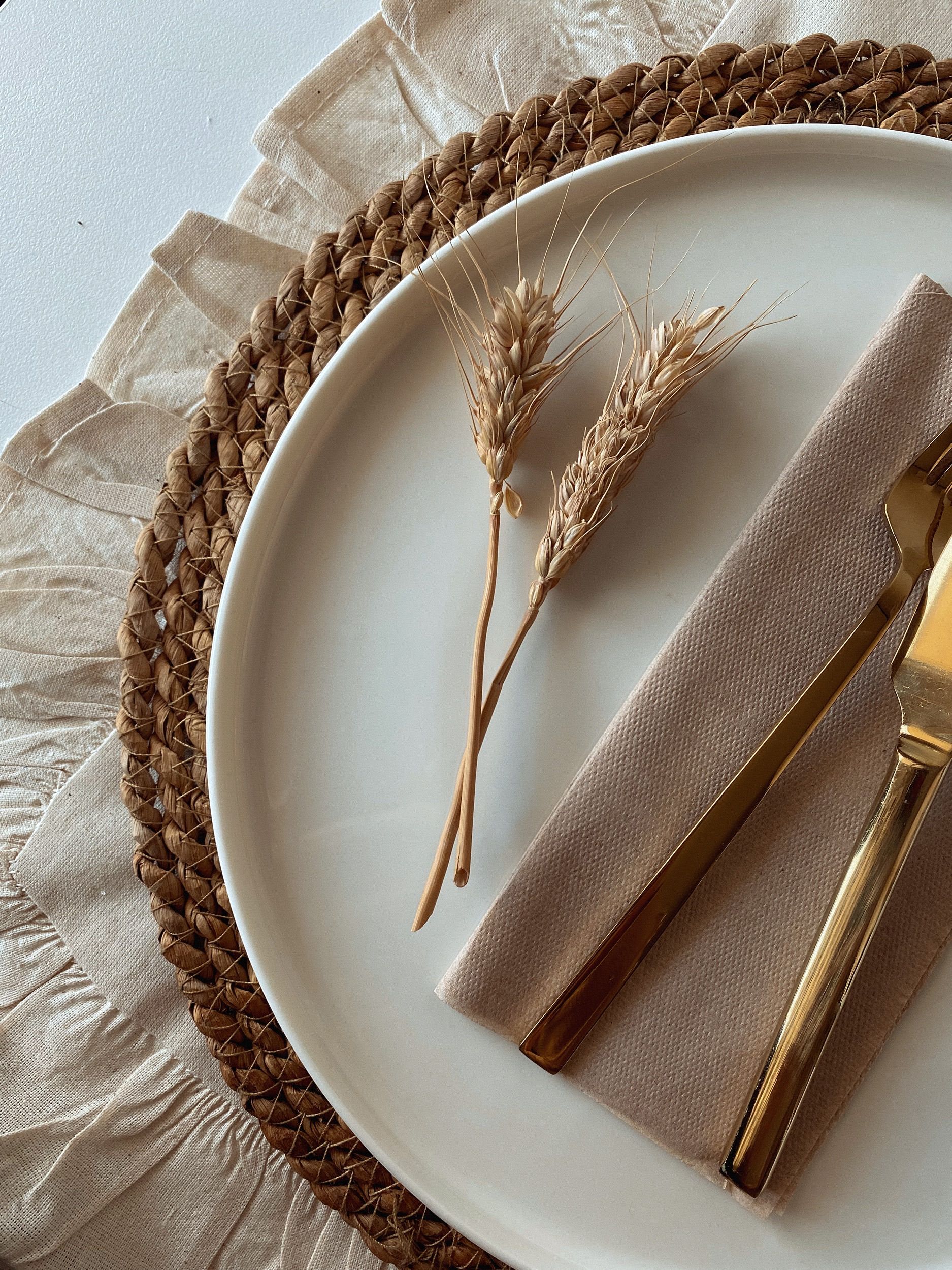 Pale plate on a woven placemat with beige fabrics, wheat stems and gold-toned cutlery