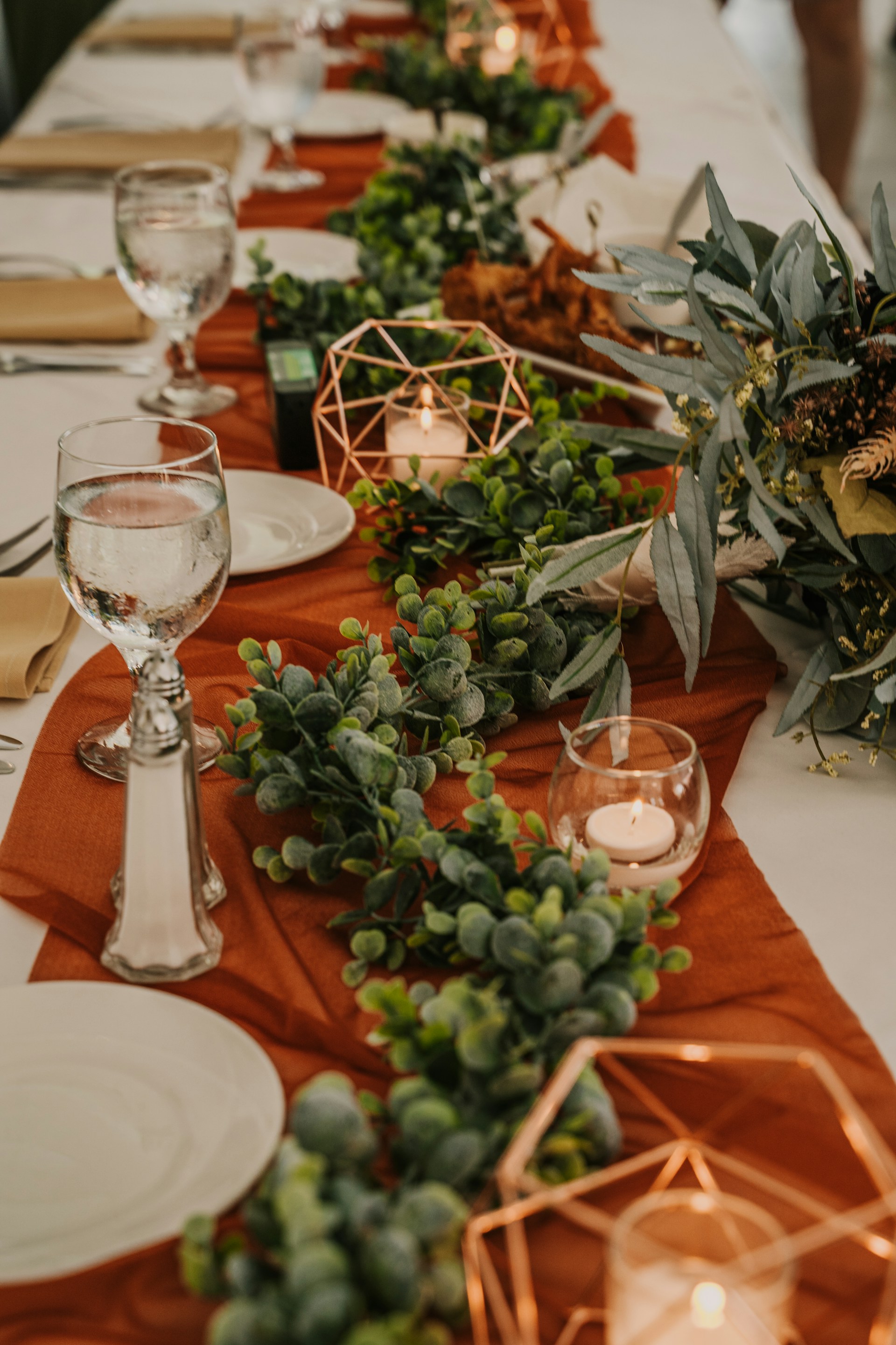 Dining table with a terracotta runner, greenery, pale plates, clear glassware and small metal candle holders