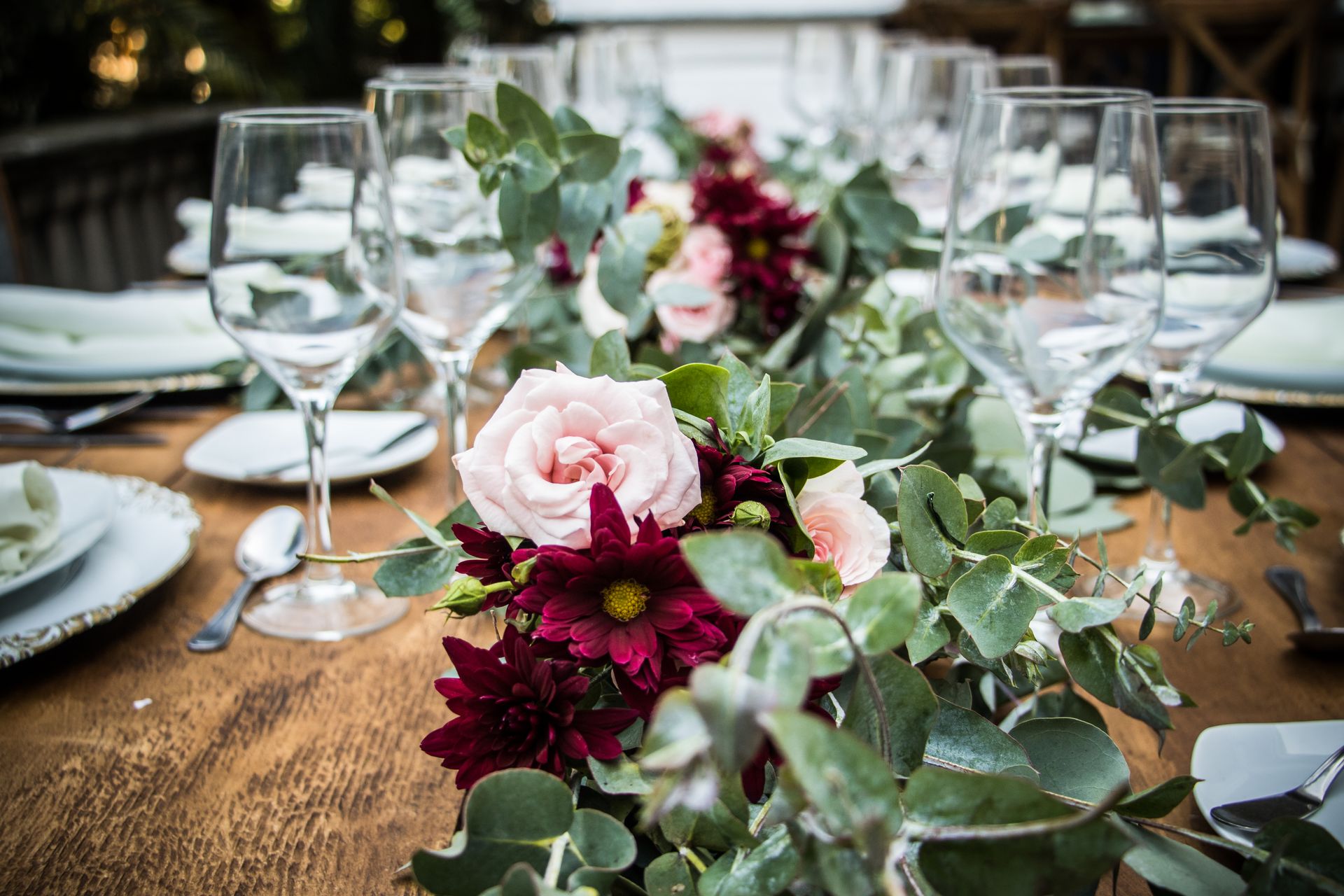 Wooden dining table with burgundy flowers, pale pink roses, greenery, white plates and clear wine glasses